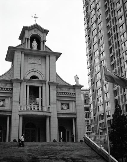 Ilford HP5 Plus 400 (6x7)

English
A black-and-white photograph of a church facade with classical architecture. The building features a central entrance with a triangular pediment, columns, and statues of religious figures. Above the entrance, there is a cross at the top. The church is flanked by tall, modern high-rise buildings. A flagpole with a flag is visible on the right side.
中文
一张黑白照片，展示了一座具有古典建筑风格的教堂正面。建筑物中间有一个三角形山墙的入口，两侧有柱子和宗教人物的雕像。入口上方顶部有一个十字架。教堂两侧是高大的现代高层建筑。右侧可以看到一根旗杆和一面旗帜。