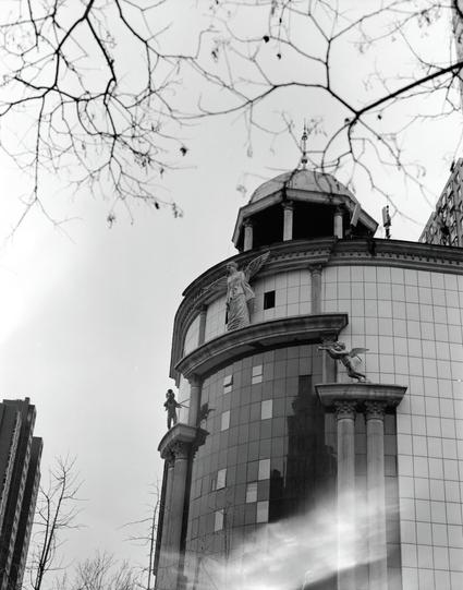 Ilford HP5 Plus 400 (6x7)

English
A black-and-white photograph of a cylindrical building with a domed roof. The structure features classical architectural elements, including columns and statues of angels. The building is partially obscured by bare tree branches in the foreground. The sky is clear, and the image captures the contrast between the classical and modern elements in the background.
中文
一张黑白照片，展示了一座带有圆顶屋顶的圆柱形建筑。建筑物具有古典建筑元素，包括柱子和天使雕像。建筑物部分被前景中光秃秃的树枝遮挡。天空晴朗，图像捕捉了古典与现代元素之间的对比。