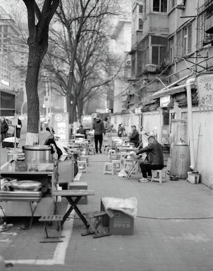Ilford HP5 Plus 400 (6x7)

English
A black-and-white photograph of a bustling street. The scene includes several food stalls with tables, chairs, and cooking equipment. A man is seated at a table on the right, while other people are walking or standing in the background. Bare trees line the street, and buildings with balconies and signs in Chinese characters are visible.
中文
一张黑白照片，展示了一个热闹的街头。场景中有几个食品摊位，摆放着桌子、椅子和烹饪设备。右侧有一名男子坐在桌子旁，其他人在背景中行走或站立。光秃秃的树木排列在街道两侧，可以看到带有阳台和中文字标志的建筑。