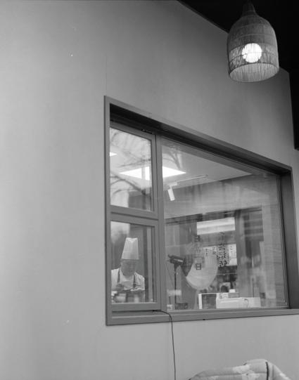 Ilford HP5 Plus 400 (6x7)

English
A black-and-white photograph showing a chef in a white hat and apron, visible through a rectangular window. The chef appears to be working in a kitchen. Above the window, a hanging light fixture with a cylindrical shade is mounted on the wall. The window reflects some indoor elements, including a sign with Chinese characters.
中文
一张黑白照片，通过一个矩形窗户可以看到一位戴白色厨师帽和围裙的厨师。厨师看起来正在厨房工作。窗户上方的墙上悬挂着一个带圆柱形灯罩的灯具。窗户反射出一些室内元素，包括一个写有中文字的标志。