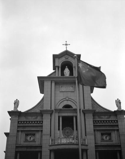 Ilford HP5 Plus 400 (6x7)

##English
Black and white photograph of a church facade. The building features a central tower with a cross at the top. A statue of a robed figure stands in a niche above the main entrance. Flanking the entrance are two statues on pedestals. Above the entrance, Chinese characters are visible. Two clocks are embedded in the facade on either side of the entrance. A Chinese flag flying on a flagpole to the front of the building.
##中文
黑白照片，展示一座教堂的正立面。建筑中央有一座塔楼，顶部立有十字架。主入口…