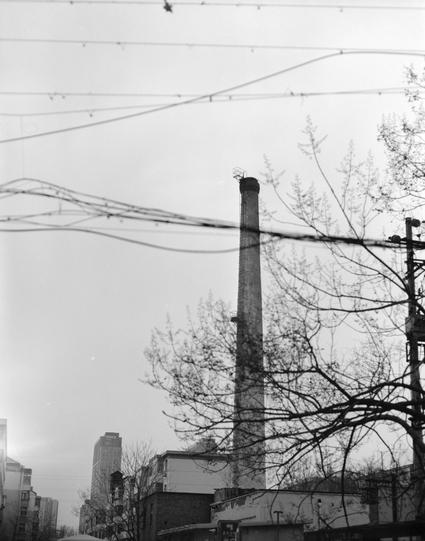 Ilford HP5 Plus 400 (6x7)

##English
Black and white photograph of an urban scene. A tall, cylindrical smokestack rises above a cluster of low-rise buildings with bare trees in the foreground. Overhead, a network of power lines crisscrosses the sky. In the background, a tall modern skyscraper is partially visible through the haze.
##中文
黑白照片，展示城市景观。一根高大的圆柱形烟囱耸立在一群低层建筑之上，前景是光秃秃的树木。上方是交错的电线网络。背景中，一座高大的现代摩天大楼在雾霭中若隐若现。