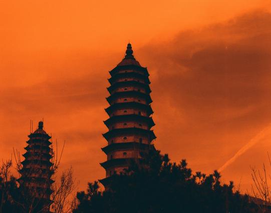 Harman Red 125 (6x7)

##English
Silhouette of two multi-tiered pagodas against a bright orange sky. The pagodas, with their curved eaves, rise above a line of dark trees. The sky is mostly clear with a few scattered clouds, and the sun casts a warm glow over the scene.
##中文
两座多层宝塔在明亮的橙色天空下的剪影。宝塔的弯曲屋檐高耸于一排深色树木之上。天空大部分晴朗，散布着几朵云彩，太阳为整个场景洒下温暖的光芒。