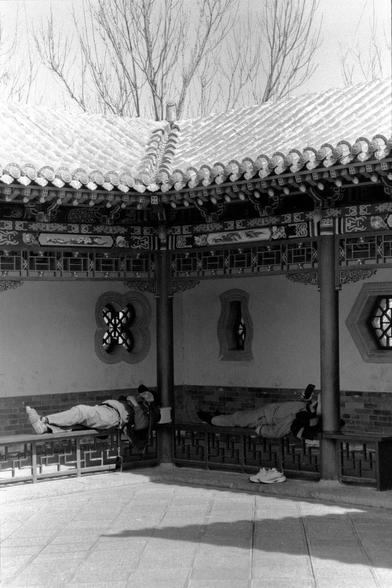 Lucky LUCKY SHD 400 (FF)

##English
Black-and-white photograph of a traditional Chinese building with ornate wooden eaves and a tiled roof. Two individuals are lying down on a wooden bench under the eaves, resting. The wall behind them features intricate geometric and floral lattice windows. Bare trees are visible in the background, suggesting a winter or early spring setting.
##中文
黑白照片，展示了一座传统中式建筑，建筑屋檐木雕精美，屋顶铺有瓦片。两个人躺在屋檐下的木长椅上休息。他们身后的墙上有精美的几何和花卉图案的格子窗。背景中可见光秃秃的树木，暗示这是冬季或早春时节。