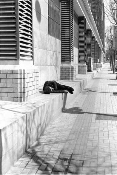 Lucky LUCKY SHD 400 (FF)

##English
Black-and-white photograph of a modern building with horizontal louvered windows. A person is lying down on a series of wide, low concrete steps leading to the building. The sidewalk is paved with light-colored tiles, and the scene is bathed in sunlight, casting distinct shadows.
##中文
黑白照片，拍摄的是一栋现代建筑，窗户为水平百叶式。一个人躺在通往建筑的宽阔低矮的水泥台阶上。人行道铺有浅色瓷砖，阳光明媚，投射出清晰的影子。