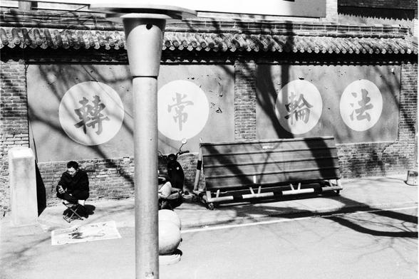 Lucky LUCKY SHD 400 (FF)

##English
Black-and-white photograph of a traditional Chinese building with a tiled roof. The wall features four large circular signs with Chinese characters: "轉" (turn), "常" (often), "輪" (wheel), and "法" (law). In front of the building, two individuals are seated on the ground, one on a small stool, engaged in calligraphy or painting on large sheets of paper. A wooden bench and a tall lamp post are also visible in the foreground.
##中文
黑白照片，拍摄的是一栋传统中式建筑，屋顶为瓦片结构。墙上挂有四个大…