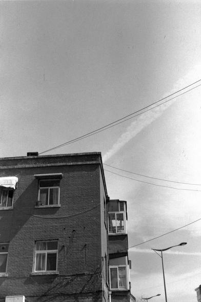 Lucky LUCKY SHD 400 (FF)

##English
Black-and-white photograph of the upper portion of a brick building. The building has multiple windows with white frames. Electrical wires stretch across the top of the image. The sky is clear, with a contrail visible in the upper right corner.
##中文
黑白照片，展示了一栋砖楼的上部。楼上有多个带白色框架的窗户。电线横跨在图片的上方。天空晴朗，右上角有一条飞机凝结尾迹。