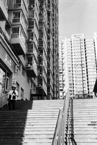 Lucky LUCKY SHD 400 (FF)

##English
A black-and-white photograph of a tall, modern apartment building with numerous windows and balconies. A person wearing a hat and carrying a bag is walking down a set of wide, concrete stairs in the foreground. The stairs are flanked by metal railings. The building has visible air conditioning units attached to its exterior. The image captures the urban environment and the contrast between the person and the towering structure.
##中文
一张黑白照片，展示了一幢高大的现代公寓楼，楼体有众多…