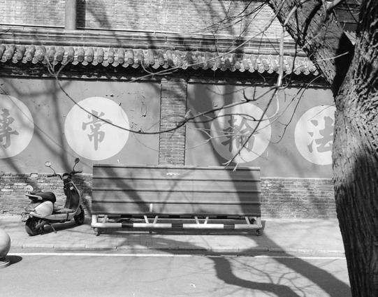 Ilford HP5 Plus 400 (6x7)

English
A black-and-white photograph of a traditional Chinese building with a tiled roof featuring decorative eaves. The wall has four large circular signs with Chinese characters: "转" (turn), "常" (often), "轮" (wheel), and "法" (law/method). A wooden bench is positioned in front of the wall, and a scooter is parked to the left of the bench. Bare tree branches cast shadows on the wall and ground.
中文
一张黑白照片，拍摄的是一座传统中式建筑，屋顶为瓦片结构，屋檐装饰精美。墙上有四个大型圆形标志，分别写着汉字：“轉”、“常”、“輪”、“法”。墙…