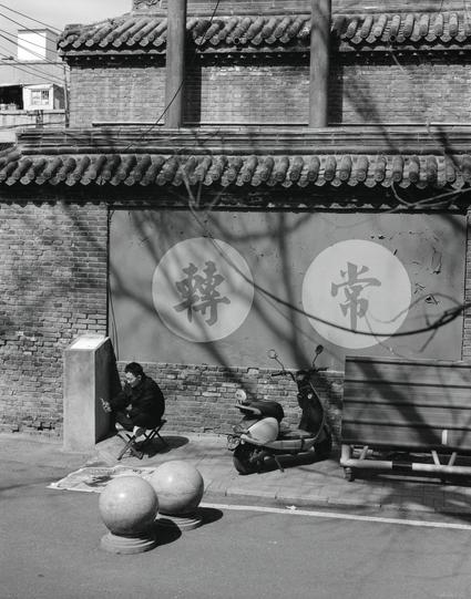 Ilford HP5 Plus 400 (6x7)

English
A black-and-white photograph of a traditional Chinese building with a tiled roof and decorative eaves. The wall features two large circular signs with Chinese characters: "转" (turn) and "常" (often). A person is seated on a folding chair in front of the wall, using a mobile phone. A scooter is parked nearby, and two large spherical stone decorations are placed on the ground. Bare tree branches and electrical wires are visible.
中文
一张黑白照片，拍摄的是一座传统中式建筑，屋顶为瓦片结构，屋檐装…