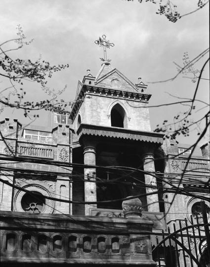 Ilford HP5 Plus 400 (6x7)

##English
Black-and-white photograph of a Gothic-style church building. The structure features a tall, pointed tower with a cross at the top, arched windows, and intricate stone carvings. The facade includes decorative columns, circular rose windows, and a balcony with a scalloped edge. Bare tree branches and power lines partially obscure the view.
##中文
黑白照片，展示一座哥特式教堂建筑。建筑特色包括一个高耸的尖塔，塔顶有一个十字架，拱形窗户以及精美的石雕装饰。立面有装饰性柱子、圆形玫瑰窗和一个带有锯齿边缘的阳台。裸露的树枝和电线部分遮挡了视线。