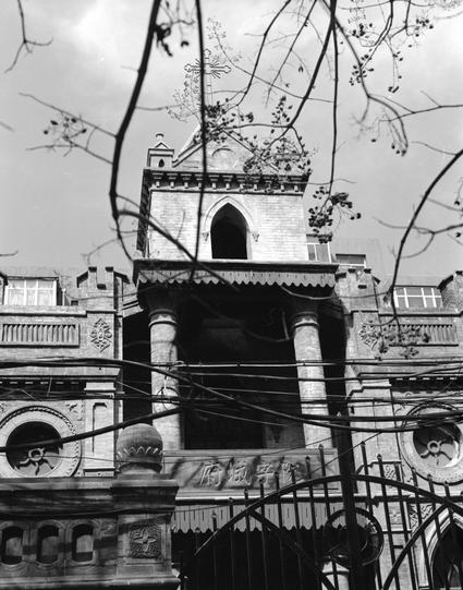 Ilford HP5 Plus 400 (6x7)

##English
Black-and-white photograph of the same Gothic-style church building from a slightly different angle. The tower with a cross at the top is visible, along with arched windows, decorative columns, and circular rose windows. The facade features Chinese characters: "府城字院" . Bare tree branches and power lines partially obscure the view.
##中文
黑白照片，从稍微不同的角度展示同一座哥特式教堂建筑。可见带有十字架的塔顶、拱形窗户、装饰性柱子和圆形玫瑰窗。立面上有中文字样：“府城字院”。裸露的树枝和电线部分遮挡了视线。