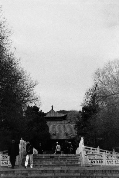 Kentmere PAN 400 (FF)

English
A black-and-white photograph of a traditional East Asian building with a tiled roof, partially obscured by bare trees. A group of people is walking on a stone bridge with ornate railings, leading toward the building. The scene is set in a serene, natural environment with trees on either side.
中文
一张黑白照片，拍摄的是一座传统东亚建筑，其瓦片屋顶被光秃的树木部分遮挡。一群人正在一座装饰精美的石桥上行走，朝建筑方向前进。场景位于一个宁静的自然环境中，两侧种满了树木。
