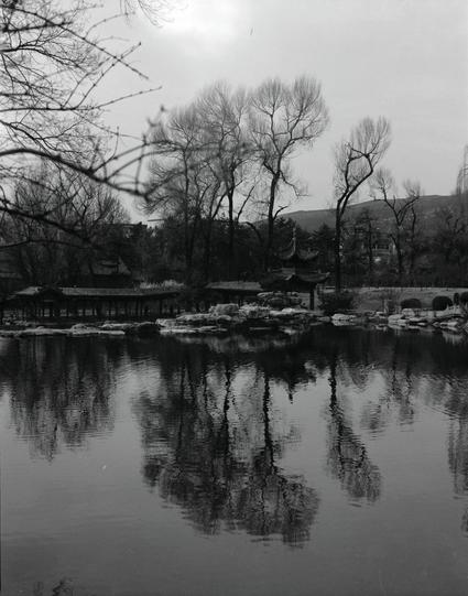 Ilford HP5 Plus 400 (6x7)

English
A black-and-white photograph of a tranquil park scene. A calm lake dominates the foreground, reflecting leafless trees and traditional Chinese pavilions with curved, tiled roofs. The bare trees, likely in winter or early spring, stand tall around the lake. In the distance, a hill with sparse vegetation adds depth to the serene landscape.
中文
一张黑白照片，展现了宁静的公园景色。前景是一片平静的湖水，倒映着光秃的树木和传统中式凉亭的弯曲瓦顶。光秃的树木可能处于冬季或早春，环绕在湖泊周围。远处，一个稀疏植被的小山为宁静的景观增添了深度。