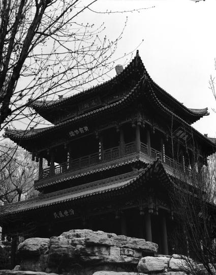Ilford HP5 Plus 400 (6x7)

English
A black-and-white photograph of a two-story traditional Chinese pavilion. The pavilion has a tiled roof, ornate wooden details, and is elevated on stone pillars. Chinese characters are visible on the signage hanging from the structure. Bare tree branches frame the top of the image, and rocky elements are visible at the base of the pavilion.
中文
一张黑白照片，展示了一座两层的传统中式凉亭。凉亭有瓦片屋顶、精美的木质装饰，并建在石柱上。悬挂的牌匾上有中文字样。光秃的树枝构成了图片的上框，凉亭底部可见岩石元素。