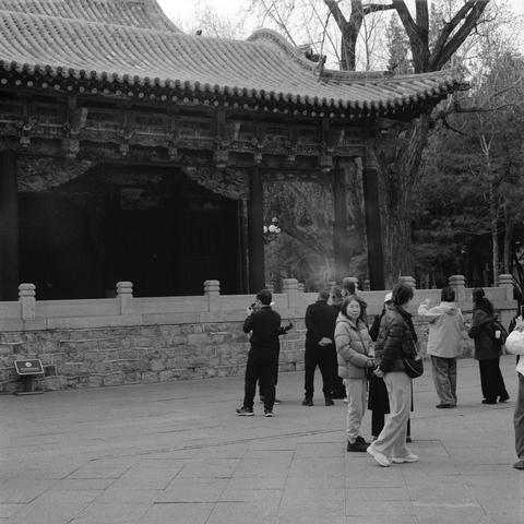 Ilford HP5 Plus 400 (6x7)

English
A black-and-white photograph of a group of people standing in front of a traditional Chinese building. The building features intricate wooden architecture with a tiled roof and a stone foundation. The people, dressed in casual winter clothing, are engaged in conversation or taking photos. Bare trees surround the area, and a stone railing is visible in the foreground.
中文
一张黑白照片，一群人站在一座传统中式建筑前。建筑具有精美的木质结构、瓦片屋顶和石头地基。人们身着休闲冬装，正在交谈或拍照。周围是光秃的树木，前景可见一道石栏杆。