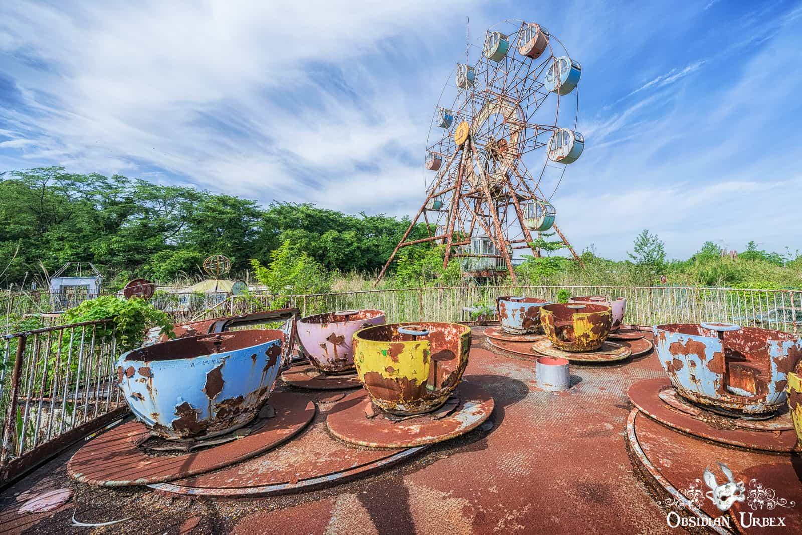 An abandoned Japanese theme park features a rusty tea cup ride in the foreground and a dilapidated Ferris wheel