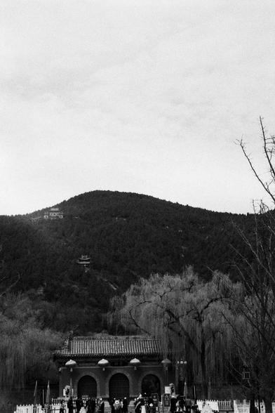 Kentmere PAN 400 (FF)

English
Black-and-white photo of a traditional Chinese building with a multi-tiered roof, set against a forested hillside. The building has arched doorways and is surrounded by bare trees. The hill in the background is densely covered with trees, and a small structure is visible at the top of the hill.
中文
黑白照片，展示一座传统中式建筑，多层屋顶，背靠森林覆盖的山坡。建筑有拱形门，周围是光秃秃的树木。后方的山坡上密布树木，山顶上有一个小建筑。