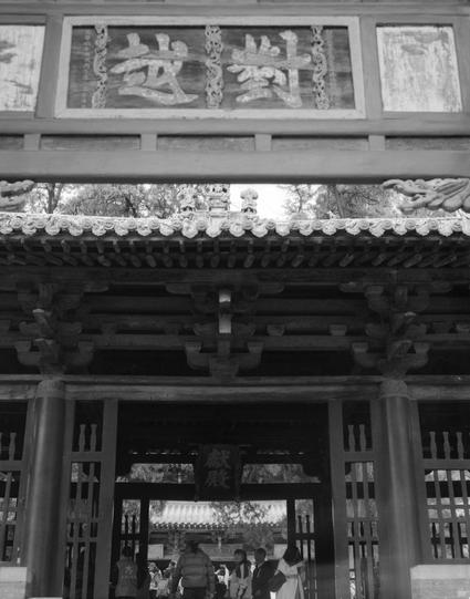 Kentmere PAN 400 (6x7)

English
Black-and-white photo of a traditional Chinese gate with intricate wooden carvings. The gate features a large rectangular signboard at the top with Chinese characters. Below the signboard, there are decorative carvings of dragons and geometric patterns. The gate has multiple layers of tiled roofs with upturned eaves. People are visible walking through the gate in the background.
中文
黑白照片，展示一个传统中式城门，门上有精美的木雕。城门顶部有一块巨大的长方形匾额，上面刻有中文字。匾额下方是龙和几何图案的装饰雕刻。城门有多层瓦顶，屋檐向上翘起。背…
