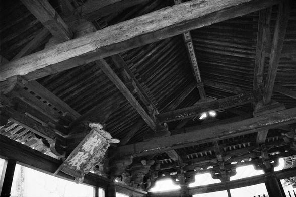 Kentmere PAN 400 (FF)

##English
Black-and-white photograph of the interior of a traditional Chinese wooden structure. The image shows a high, intricately carved wooden ceiling with exposed beams and decorative elements. The ceiling appears aged, with visible wear and patina on the wood. Light filters through gaps in the roof, illuminating parts of the interior.
##中文
黑白照片，拍摄的是一座传统中式木结构建筑的内部。图中展示了高耸、精美雕刻的木质天花板，露出的梁木和装饰性元素清晰可见。天花板看起来有年头了，木头上有明显的磨损和光泽。光线从屋顶的缝隙中透过，照亮了内部的部分区域。