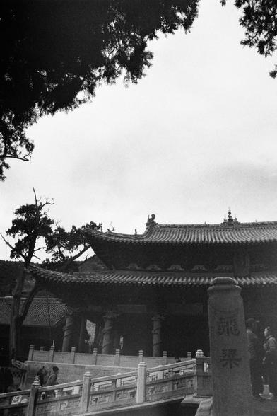 Kentmere PAN 400 (FF)

##English
Black-and-white photograph of a traditional Chinese temple complex. The image captures a stone bridge with carved railings leading to the entrance of the temple. The temple roof is tiled with upturned eaves and decorative elements. A tall stone pillar with Chinese characters is visible on the right. Trees frame the scene, and a few people are walking near the entrance.
##中文
黑白照片，拍摄的是一座传统中式寺庙建筑群。图中有一座石桥，桥上雕刻有精美的栏杆，通向寺庙的入口。寺庙屋顶为瓦片结构，屋檐向上翘起，并有装饰性元素。右侧可见一根高大的石柱，上面刻有…
