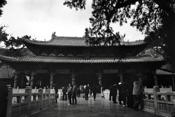 Kentmere PAN 400 (FF)

##English
Black-and-white photograph of a traditional Chinese temple entrance. The scene features a large, ornate building with a multi-tiered, curved roof adorned with intricate eaves. The structure is supported by tall, carved stone pillars. In front of the temple, a group of people stands and walks on a wide, stone-paved pathway. The pathway is flanked by low, decorative stone railings. Tall trees with bare branches frame the top of the image, adding depth and contrast…