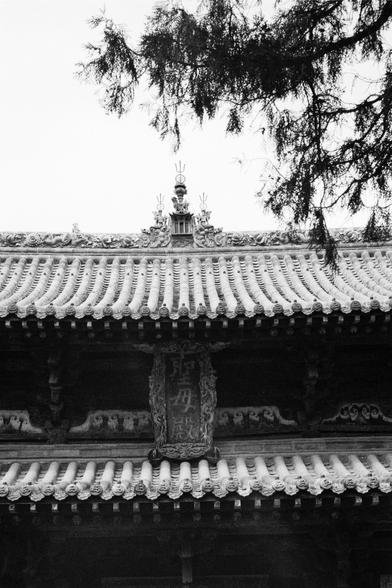 Kentmere PAN 400 (FF)

##English
Black-and-white close-up of the roof of a traditional Chinese building. The roof is highly detailed, with multiple layers of curved tiles and ornate carvings. At the peak, there is a decorative ridge with intricate designs, including a small pavilion-like structure. The eaves are adorned with elaborate patterns, and a vertical plaque with Chinese characters is visible in the center.
##中文
黑白照片，特写镜头拍摄的是一座传统中国建筑的屋顶。屋顶装饰精美，有多层弯曲的瓦片和精美的雕刻。屋脊处有一个装饰性的脊饰，包含一个小亭子状的结构。屋檐饰…