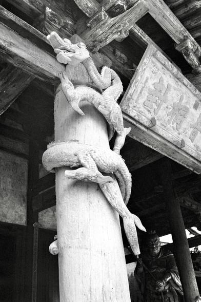 Kentmere PAN 400 (FF)

##English
Black-and-white photograph of a carved stone pillar in front of a traditional Chinese building. The pillar is wrapped by a detailed stone sculpture of a dragon, its body coiled around the column. The dragon's head is positioned at the top, facing outward. The building behind the pillar has a wooden structure with visible beams and a tiled roof. A person stands partially visible in the background.
##中文
黑白照片，拍摄的是一根位于传统中国建筑前的雕刻石柱。石柱上缠绕着一个详细的石龙雕塑，龙身环绕在柱子上，龙头位于顶部，面向外…