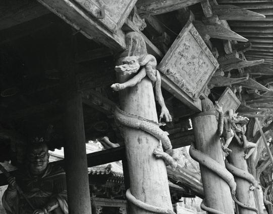 Kentmere PAN 400 (6x7)

##English
Black-and-white photograph of a traditional wooden structure with intricate carvings. Two tall, cylindrical wooden pillars are wrapped by sculpted dragons, their bodies coiling around the columns. Above, a wooden sign with Chinese characters is partially visible. The roof beams and eaves are dark and weathered, adding to the aged appearance of the building.
##中文
黑白照片，展示一个传统木结构建筑，雕刻精美。两根高大的圆形木柱被雕刻的龙环绕，龙的身体缠绕在柱子上。上方有一块木制的中文字牌匾，部分可见。屋顶的梁和屋檐颜色深暗，显示出建筑的古老感。