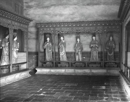 Kentmere PAN 400 (6x7)

##English
Black-and-white photograph of a traditional interior with a raised wooden platform. Five statues of robed figures stand in a row on the platform, each with distinct postures and attire. The backdrop features intricate carvings and patterns. The floor is tiled, and the walls are plain, emphasizing the statues and the platform.
##中文
黑白照片，展示一个传统室内空间，中央有一个高起的木质平台。平台上并排站着五尊穿着长袍的人物雕像，每尊雕像姿态和装束各不相同。背景是精美的雕刻和图案。地面是瓷砖铺设，墙壁简洁，突出了雕像和平台。