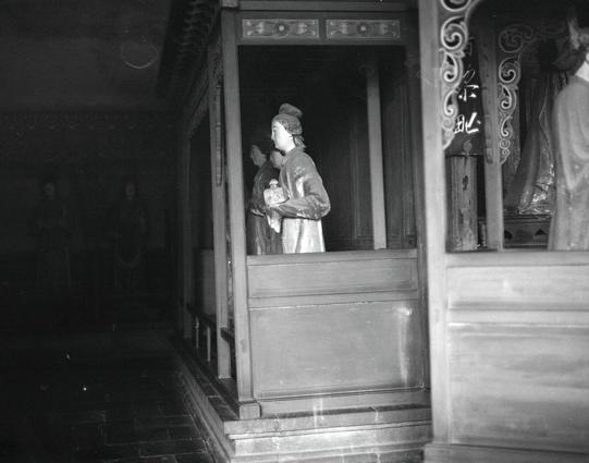 Kentmere PAN 400 (6x7)

##English
Black-and-white photograph of a dimly lit interior space. A wooden statue of a robed figure holding an object stands in a doorway, framed by ornate carvings. The doorway is part of a larger wooden structure with decorative elements. The lighting is low, creating a shadowy atmosphere.
##中文
黑白照片，展示一个光线昏暗的室内空间。一个穿着长袍、手持物品的木雕人物站在门口，门框饰有精美的雕刻。门口是更大的木结构的一部分，装饰元素丰富。光线较暗，营造出一种阴影笼罩的氛围。