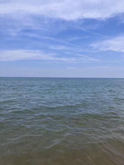 Standing in the shallow surf, looking north into the Atlantic Ocean. The sky is partly cloudy, but revealing a bright blue sky behind. The water is mostly calm, ranging in color from greenish blue to a greenish brown. 