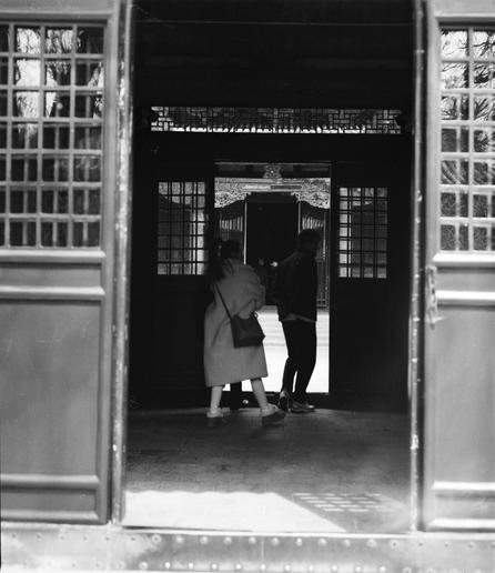 Kentmere PAN 400 (6x7)

English
A black-and-white photograph showing the entrance of a traditional Chinese building. Two individuals stand inside the doorway, which is framed by open wooden doors with geometric lattice patterns. The doors lead to a courtyard with a tiled floor. The scene is dimly lit, with light filtering through the open doors.
中文
一张黑白照片，展示了中国传统建筑的入口。两个人站在门内，门框由带有几何格子图案的木门组成。门通向一个铺有瓷砖地面的庭院。场景光线昏暗，光线通过打开的门洒进来。