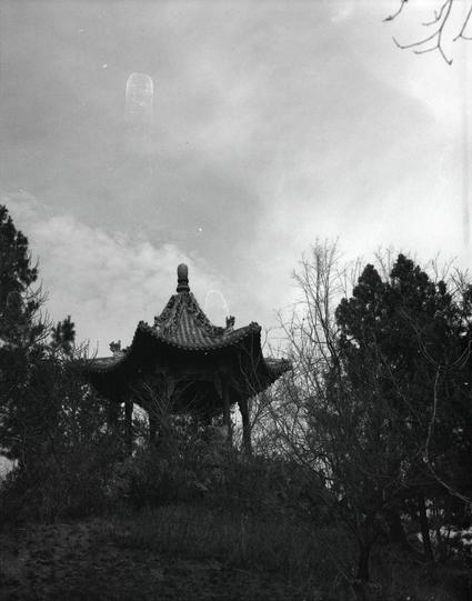 Kentmere PAN 400 (6x7)

English
A black-and-white photograph of a traditional Chinese pavilion with a tiered, ornate roof. The pavilion is surrounded by trees and shrubs, partially obscuring its base. The sky above is cloudy, with a faint circular light spot visible in the upper center of the image.
中文
一张黑白照片，拍摄的是一座中国传统亭台，屋顶为分层装饰结构。亭台被树木和灌木丛包围，部分遮挡了其基座。上方的天空多云，图片上方中央可见一个淡淡的圆形光斑。