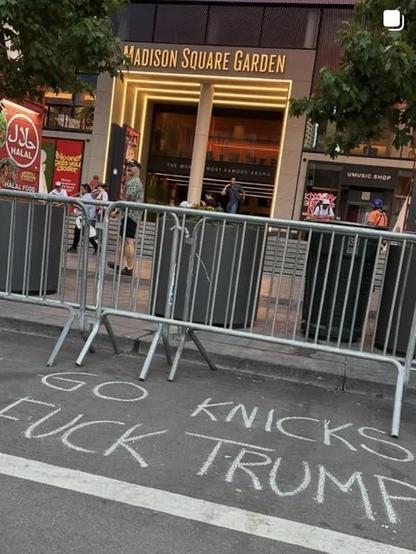 Street photo showing an entrance to Madison Square Garden, in New York City, where some sportsball is occurring tonight, at which event Trump is expected to appear. A crowd control barrier extends across the image in midground, at the bottom, scrawled in chalk, are the words, 
Go Knicks 
Fuck Trump 