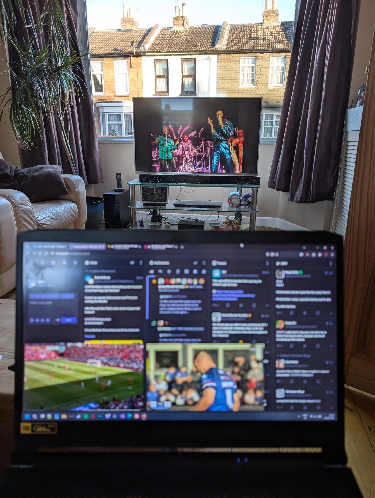 A laptop on a lap with Mastodon columns visible and two video overlays, one with football, one with Rugby League on it. In the background there's a living room with bay window and large TV showing Top of the Pops. It's bright and houses across the street can be seen.
