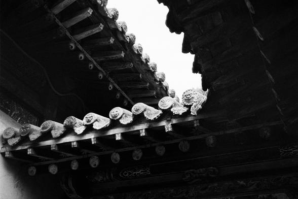 Kentmere PAN 400 (FF)

##English
Black-and-white photograph of a section of a traditional Chinese roof. The eaves are intricately carved with small, detailed figures and decorative elements. The roof tiles are layered, and the image captures the underside of the roof, showcasing the craftsmanship.
##中文
黑白照片，展示传统中式屋顶的局部。屋檐下雕刻精细，有多个小型人物和装饰性图案。屋顶瓦片层叠，画面展示了屋顶内部的精美工艺。