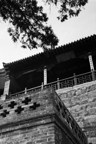 Kentmere PAN 400 (FF)

##English
Black-and-white photograph of a traditional Chinese building with a tiled roof and wooden structure. The building features a stone foundation with visible brickwork and a railing with geometric patterns. Vertical signs with Chinese characters are visible on the right side of the structure. Tree branches partially frame the top of the image.
##中文
黑白照片，展示一座传统中式建筑，屋顶为瓦片结构，主体为木质。建筑基座为石头砌成，砖石纹理清晰可见，栏杆上有几何图案。立有几块竖直的中文牌匾。画面上方有树枝部分遮挡。