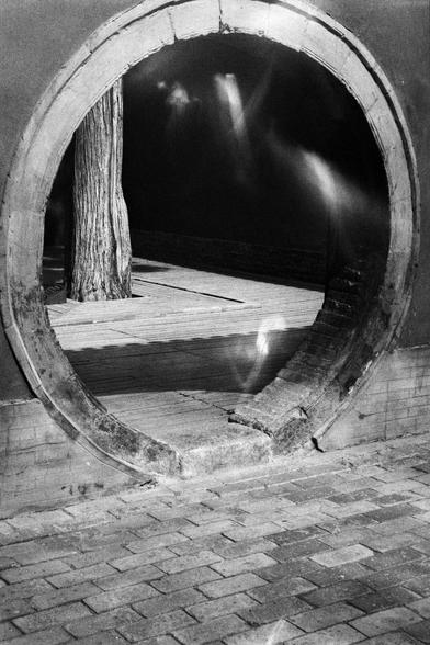 Kentmere PAN 400 (FF)

##English
Black-and-white photograph of a circular stone doorway or window, known as a moon gate. The gate is set in a brick wall, and the ground is paved with stone tiles. A tree trunk is partially visible through the opening, with blurred light and shadows in the background.
##中文
黑白照片，展示一个圆形石门（月亮门），嵌入砖墙中。地面铺有石板，门洞中可见部分树干，背景有模糊的光线和阴影。