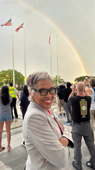 Rep Joyce Beatty, a Black woman with salt and pepper hair and a grey suit and black framed glasses, stands smiling in front of the rainbow over DC that broke out while Trump's name was being removed from the Kennedy Center. Lots of people are standing behind her taking pictures of the beautiful rainbow.