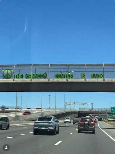 On a pedestrian bridge over a highway in Tyson's Corner, protesters have a bright green peace sign and then big green letters saying "THE KENNEDY CENTER IS BACK"