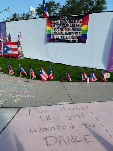 A big pride flag hangs from a wall with pictures of all the victims of the Pulse shooting in Orlando, Florida, 10 years ago. Also seen are lots of US and Puerto Rican flags. On the sidewalk it says "For those who just wanted to DANCE." Off to the left there are other slogans like "Queer lives matter" chalked in rainbow colors.