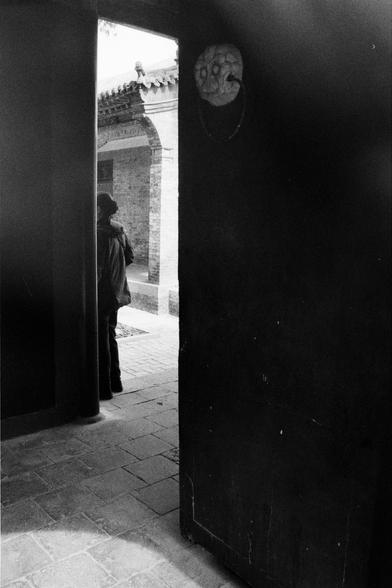 Kentmere PAN 400 (FF)

English
A black-and-white photograph showing an open wooden door with a carved circular decoration on the right side. Beyond the doorway, a person stands in a courtyard with traditional Chinese architecture, including a tiled roof and brick walls. The ground is paved with stone tiles.
中文
一张黑白照片，展示了一扇打开的木门，右侧有一个圆形雕花装饰。门外，一个人站在传统中式建筑的庭院中，建筑包括瓦屋顶和砖墙。地面铺着石板。