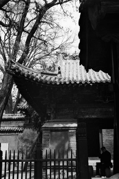 Kentmere PAN 400 (FF)

English
A black-and-white photograph of a traditional Chinese building with a tiled roof and ornate eaves. A bare tree with twisted branches stands in front of the building. A person is seated on a bench in the foreground, partially obscured by the building's shadow.
中文
一张黑白照片，展示了一座传统中式建筑，屋顶覆盖着瓦片，屋檐装饰精美。建筑前有一棵光秃的树，枝干扭曲。前景中有一个人坐在长凳上，部分被建筑的阴影遮挡。