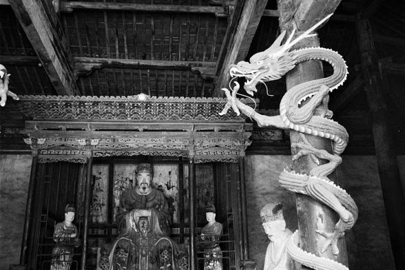 Kentmere PAN 400 (FF)

English
A black-and-white photograph of the interior of a traditional Chinese temple. A large, intricately carved wooden dragon sculpture wraps around a pillar on the right. In the background, there is a statue of a seated figure, likely a deity, surrounded by smaller statues. The ceiling features ornate wooden beams.
中文
一张黑白照片，展示了中国传统寺庙的内部。右侧有一条精美雕刻的木龙环绕在柱子上。背景中有一尊坐姿的神像，周围环绕着较小的雕像。天花板上有精美的木雕横梁。