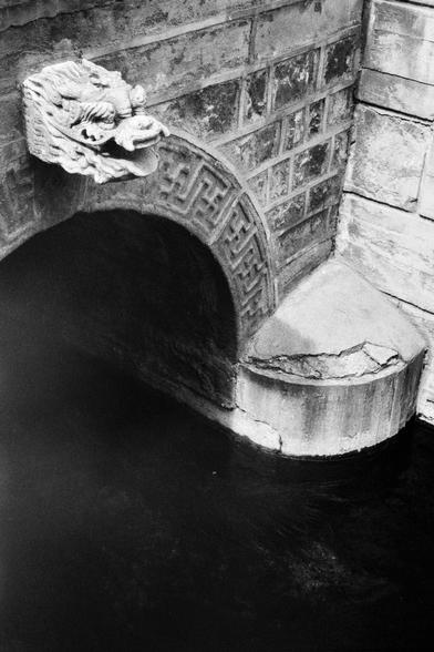 Kentmere PAN 400 (FF)

English
A black-and-white photograph of a stone archway with intricate carvings, including a dragon head at the top left. The archway is part of a brick bridge, and below it, there is a circular stone basin over the river water. The reflection of the archway is visible in the water.
中文
一张黑白照片，展示了一个石拱门，上面有精美的雕刻，左上角有一个龙头。拱门是砖桥的一部分，下方有一个圆形的石盆，在水面上。拱门的倒影清晰地映在水中。