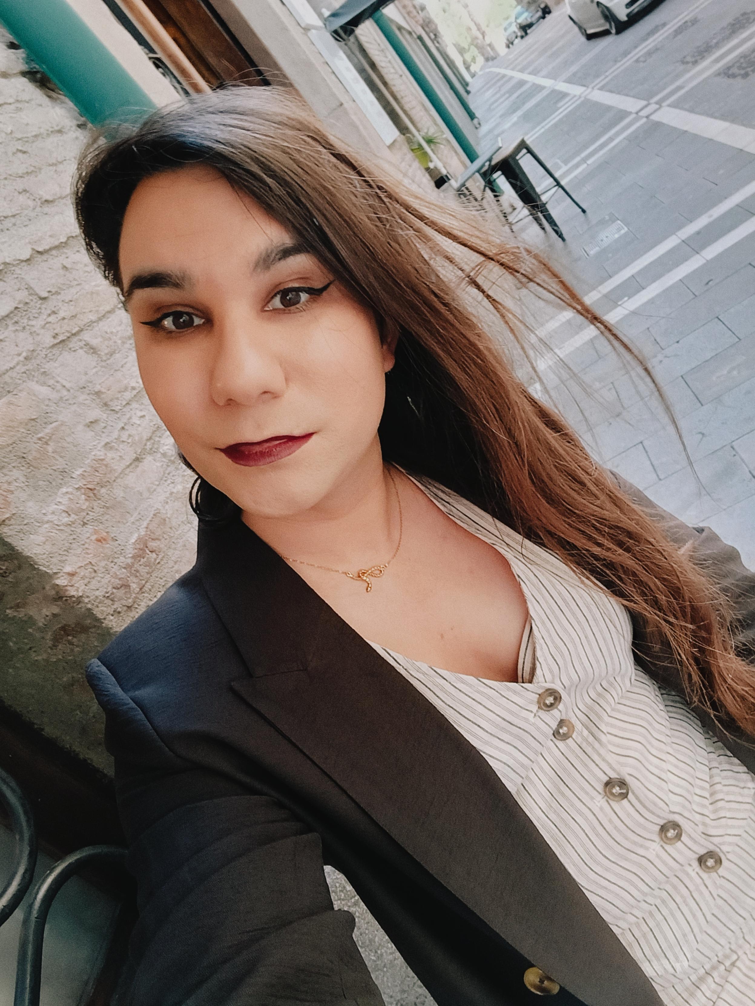 Selfie of a brunette with long hair wearing a brown suit jacket over a white buttoned shirt with thin dark vertical stripes. She's seating on a stool on a coffee shop outside on the street of a European old quarter.