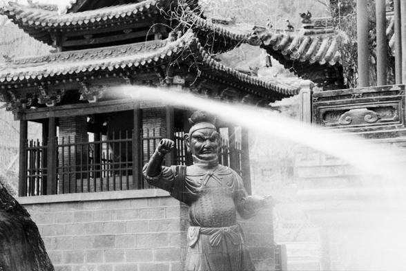 Kentmere PAN 400 (FF)

English
Black-and-white photograph of a traditional Chinese temple with intricate roof eaves. In the foreground, there is a stone statue of a muscular, fierce-looking guardian figure with a detailed headdress, armored chest, and a clenched fist raised. The background features a multi-tiered temple building with ornate wooden carvings and layered, upturned roof corners.
中文
黑白照片，拍摄的是一座传统中式寺庙，屋檐装饰精美。前景是一尊石雕守护神像，肌肉发达，面容凶猛，头戴精美头饰，身穿铠甲，一只拳头高高举起。背景是一座多层寺庙建筑，木雕精美，屋檐层叠上翘。