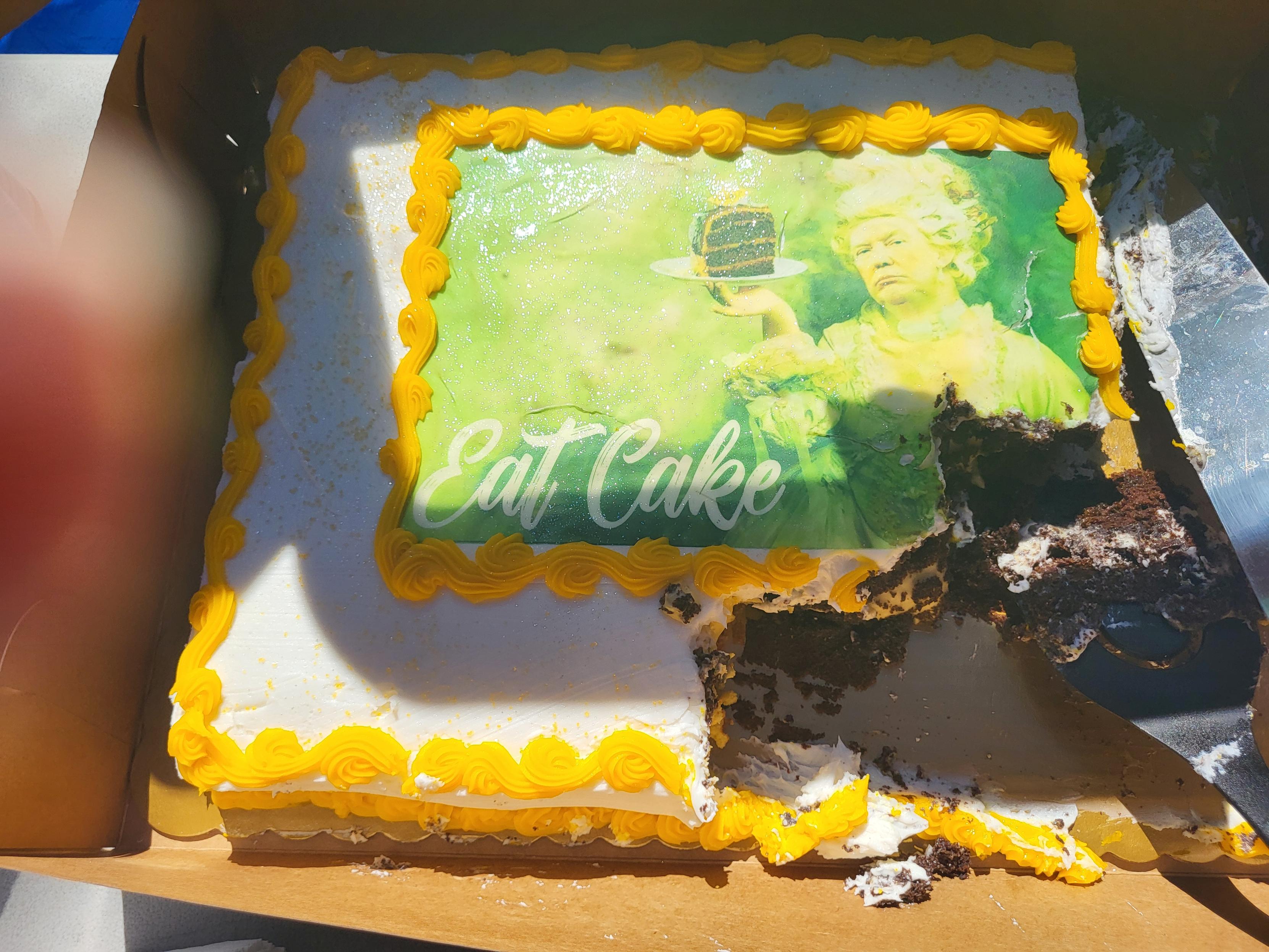 Cake at our combination protest party and mutual aid fair. A chocolate sheet cake with a cream layer and white icing, with yellow frosting scrolling around the edges. In the middle is a meme of Trump as Marie Antoinette holding a piece of chocolate cake. In white script letters it says "Eat Cake."

Also visible off to the left is my finger, because I took the picture like an idiot.