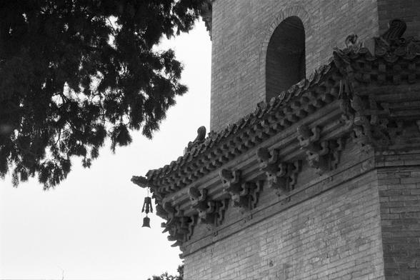 Ilford Pan 400 (FF)

English
A black-and-white close-up photograph of the eaves of a traditional Chinese building. The eaves are intricately carved with detailed patterns. A small bell hangs from the edge of the roof. Part of a tree branch is visible in the top left corner. The building is made of brick, and an arched window is partially visible.
中文
一张黑白特写照片，拍摄的是传统中国建筑的屋檐。屋檐上雕刻有精细的图案。屋顶边缘挂着一个小铃铛。左上角可见部分树枝。建筑由砖块砌成，一扇拱形窗户部分可见。